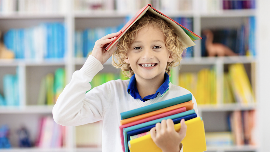 Kind hält ein aufgeschlagenes Buch wie ein Dach über den Kopf und weitere Bücher im Arm