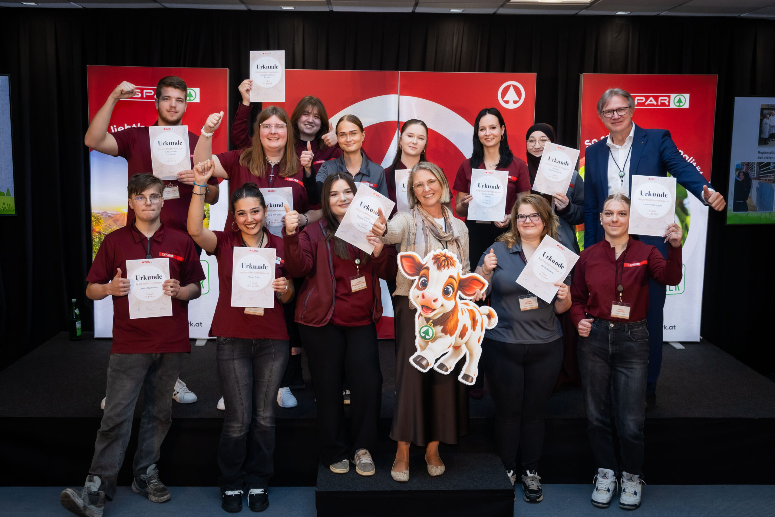 Zwölf SPAR-Lehrlinge halten stolz ihre Urkunden zur Zusatzausbildung als Regionalitätsmanager auf einer Bühne in der Hand, zusammen mit SPAR-Geschäftsführer Christoph Holzer.